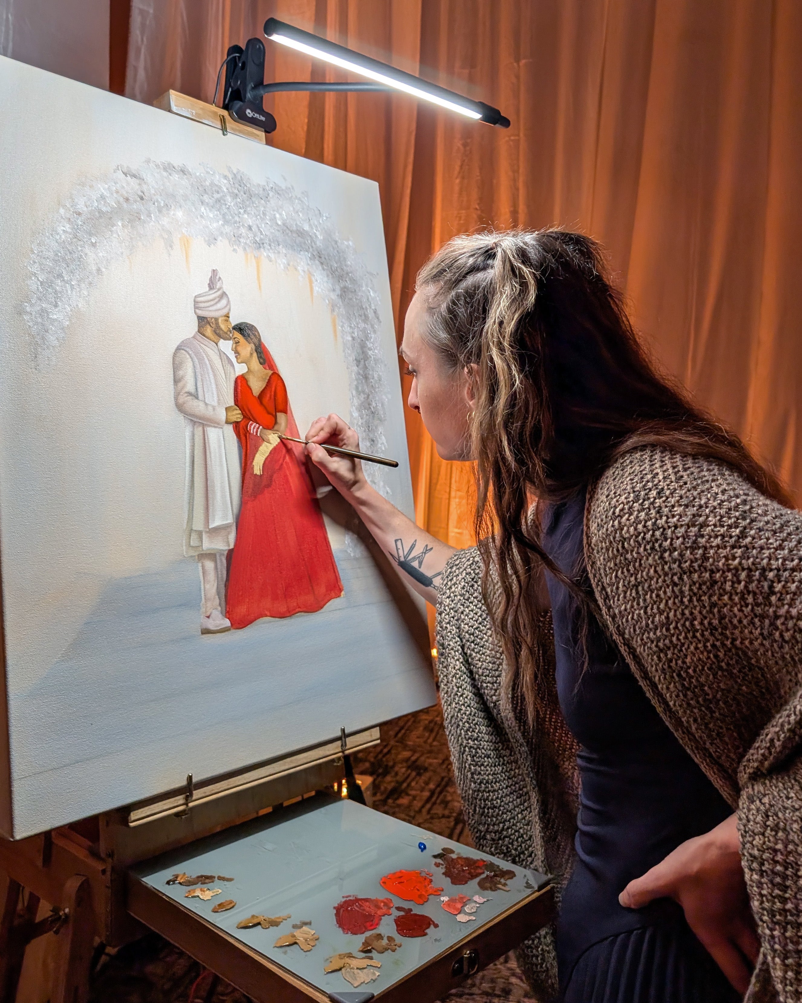 Artist painting a detailed scene of two people on an easel with a lamp illuminating the artwork.